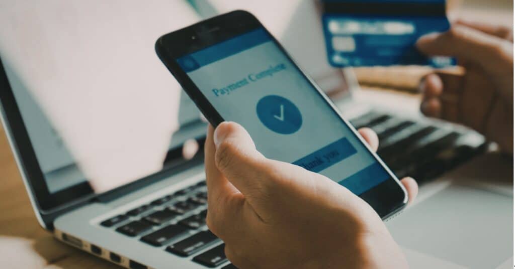 A person holds a smartphone displaying a "Payment Complete" message with a checkmark, while holding a credit card in front of an open laptop