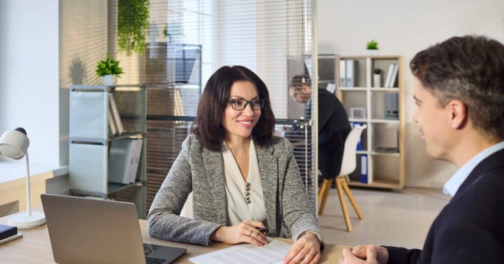 A woman in a grey blazer and glasses smiles while sitting at a desk with a laptop and papers, facing a man in a suit.