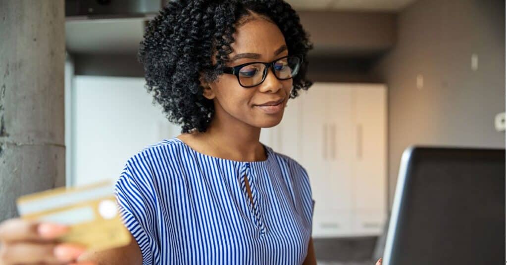 A person wearing glasses and a blue and white striped shirt holds a credit card while typing on a laptop.