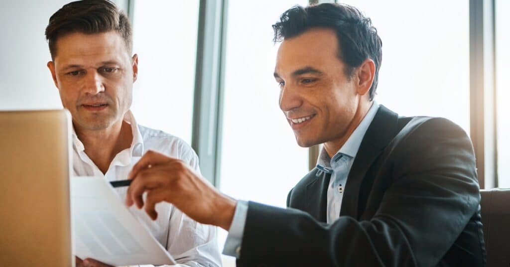 Two men in suits are looking at NCUA examination preparation papers on a desk, one is pointing at the papers with a pen and smiling.