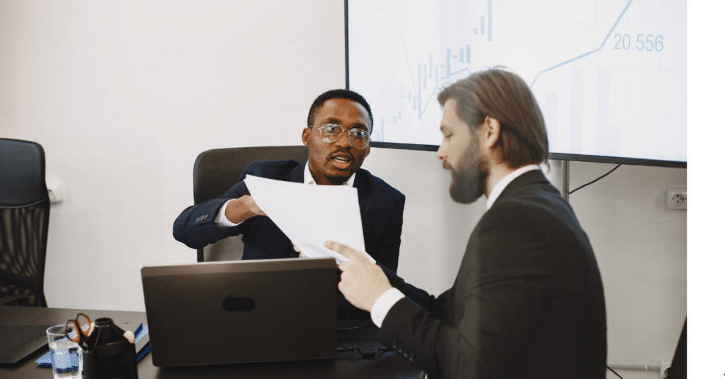 Two NCUA compliance colleagues in a meeting room gesture while talking across a conference table, with laptops and office supplies in front of them and a presentation screen showing a chart in the background.