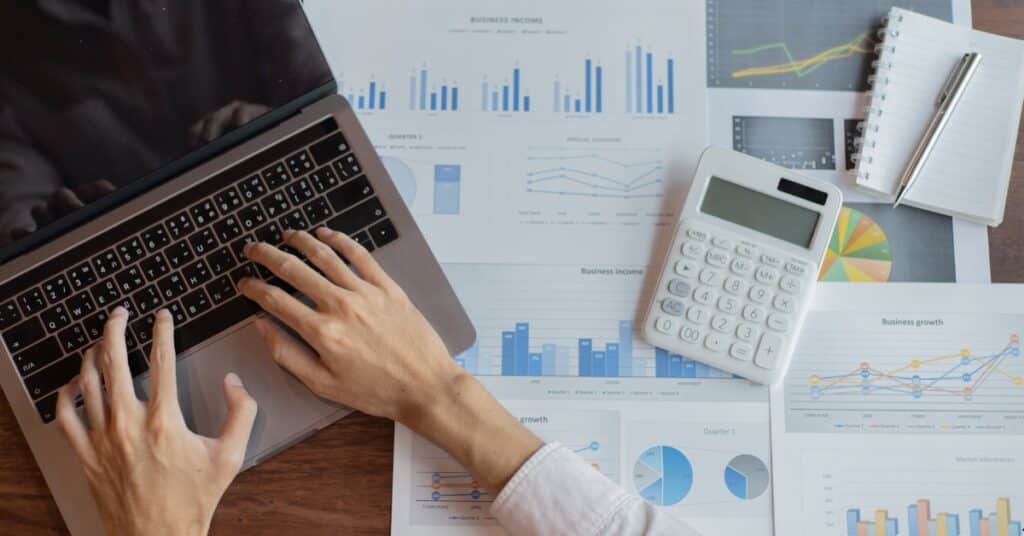A person's hands typing on a laptop surrounded by financial charts, a calculator, and a notebook with a pen.